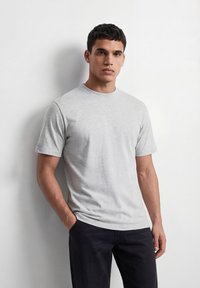 Άνδρας με κοντά σκούρα μαλλιά στέκεται μπροστά σε λευκό τοίχο, φορώντας απλό ανοιχτό γκρι κοντομάνικο μπλουζάκι και μαύρο παντελόνι, με τα χέρια στις τσέπες.