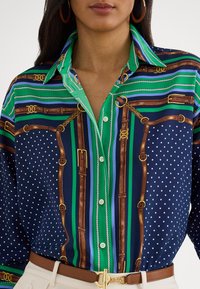 Kvinne iført en knappet flerfarget bluse med grønne, blå og brune stripete og polkaprikker mønstre, kombinert med beige bukser og et brunt belte.