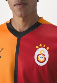Puma-trøje med et delt design: orange til venstre, rød til højre. Sort krave og detaljer, med et logo med stjerner og tekst.