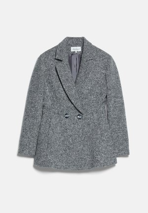 Femme portant une veste en tweed grise avec une ceinture marron, une jupe assortie, une casquette grise et des talons marron, tenant un grand sac à main marron.