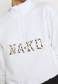 Sweatshirt blanc à fermeture éclair avec un col montant. Présente un logo "NAKD" audacieux en imprimé léopard sur le devant. Textures douces, coupe courte.