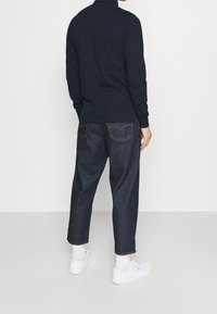 Polo de manga larga azul marino con un cuello acanalado, combinado con jeans de mezclilla oscuros de corte holgado y zapatillas blancas. Detalles de costura trasera visibles.