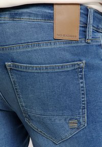 Denim jeans in medium blue, met een bruin leren taillebandlabel met "NO EXCESS" gedrukt, en kenmerkende stiksels op de achterzak.