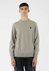 Pullover lavorato a maglia di un beige chiaro con colletto rotondo, polsini e orlo a coste, con un piccolo logo nero sul petto. Indossato con jeans scuri.