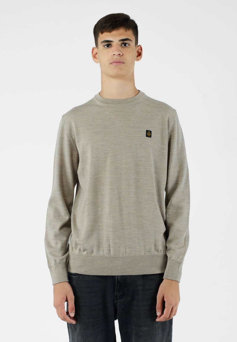 Pullover lavorato a maglia di un beige chiaro con colletto rotondo, polsini e orlo a coste, con un piccolo logo nero sul petto. Indossato con jeans scuri.