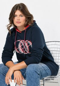 Felpa blu navy con dettagli rossi, logo in bianco e motivo rosso. Composizione in cotone misto, vestibilità comoda, abbinata a jeans, seduto su una sedia di metallo.