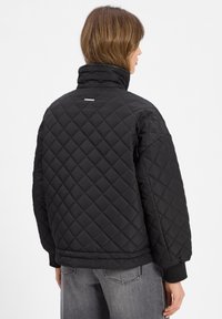 Giacca nera trapuntata con colletto alto, caratterizzata da un motivo a rombi e polsini a costine. Branding discreto sul retro.