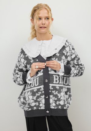 Vrouw met krullend blond haar die een wit blouse met ruches bij de kraag draagt en een grijze cardigan met bloemenpatroon en knopen.