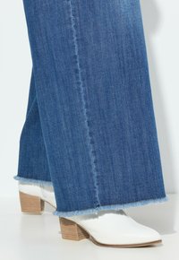 Jeans de mezclilla de tiro alto, de pierna ancha y color azul claro, con dobladillo deshilachado, combinados con botas blancas de tacón puntiagudo que cuentan con un tacón de bloque de madera.