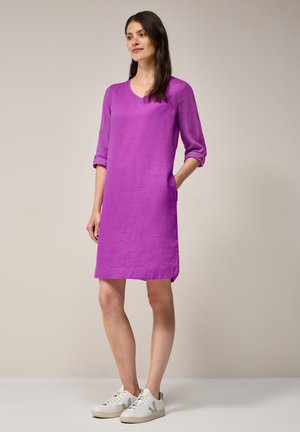 Lila Leinenkleid mit V-Ausschnitt und dreiviertellangen Ärmeln, ausgestattet mit seitlichen Taschen. Kombiniert mit weißen Sneakers.