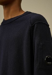 Maglione blu navy a maglia con scollatura rotonda e dettagli a coste. Presenta una tasca a toppa sulla manica con un accento di logo circolare. Texture morbida.