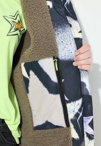 Fleecejacka med övre del i brun sherpa och mönstrad nedre del med svarta och vita stjärnor. Inkluderar en ficka med dragkedja och neonaccent.