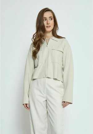 Light beige cropped jacket with a zip closure, two chest pockets, and long sleeves. Smooth texture, paired with cream trousers.