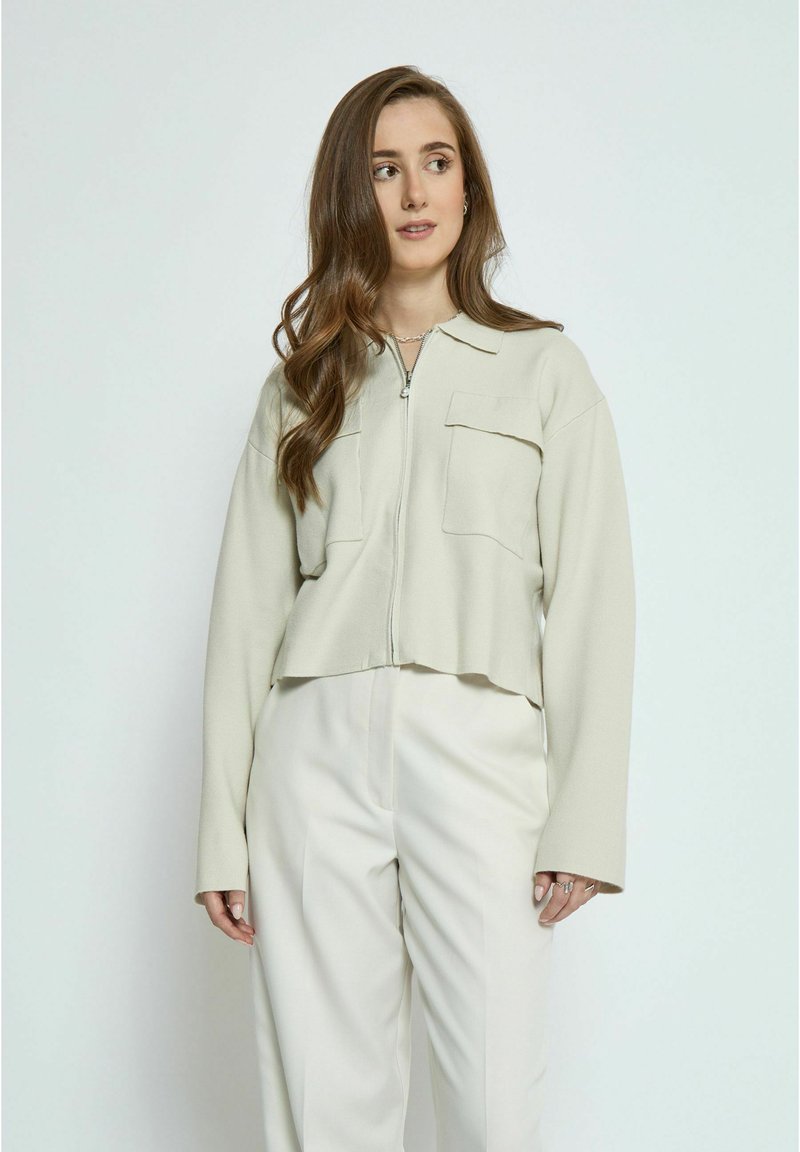 Helle beige, kurze Jacke mit Reißverschluss, zwei Brusttaschen und langen Ärmeln. Glatte Textur, kombiniert mit cremefarbenen Hosen.
