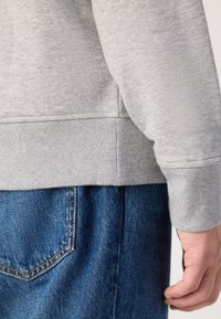 Sudadera gris con puños acanalados, mostrando una tela texturizada. Combinada con jeans de mezclilla azul con un bolsillo trasero y costuras visibles.