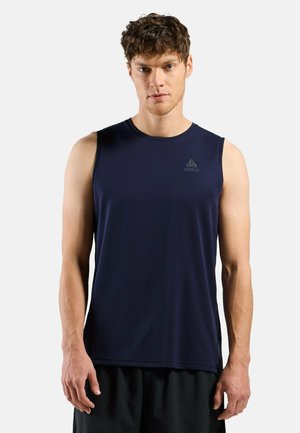 Mann trägt ein ärmelloses marineblaues Sportshirt und schwarze Shorts, steht vor einem schlichten hellen Hintergrund und schaut nach vorne.