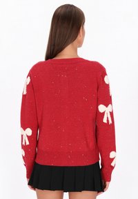 Hosszú barna hajú nő piros pulóverben, amelyen fehér masnis minta található az ujjaknál, és egy fekete, redős szoknyát visel, háttal áll.