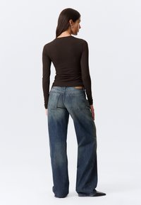 Brūns garām piedurknēm piegulošs tops un vaļējas piegriezuma izbalējuši zilie džinsi ar taisnām bikšu kājām un divām aizmugurējām kabatām, skats no muguras.
