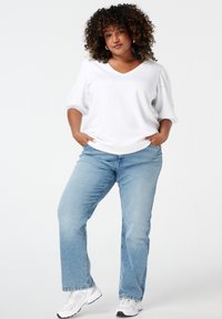 Witte pullover met een V-hals en puff mouwen, gecombineerd met lichtblauwe rechttoe rechtaan spijkerbroek en witte sportschoenen.