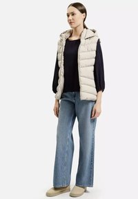 Lekki puchowy bezrękawnik w kolorze beżowym z kapturem, w połączeniu z ciemną bluzką z długim rękawem i szerokimi niebieskimi dżinsami. Obuwie to beżowe botki za kostkę.