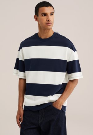Junger Mann mit kurzen dunklen Haaren, der ein marineblau-weiß horizontal gestreiftes kurzärmeliges Hemd und dunkle Hose trägt, mit einer Hand in der Tasche.