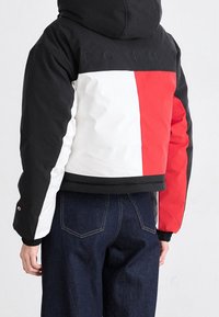 Prošívaná bunda s černou kapucí, zdobená barevnými bloky v černé, bílé a červené barvě. Texturovaná látka a vyšívané logo na zádech.