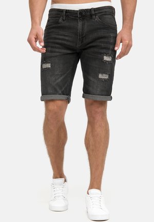 Mann trägt schwarze, hochgekrempelte, verwaschene Denim-Shorts und weiße Turnschuhe, steht mit locker an den Seiten hängenden Händen.