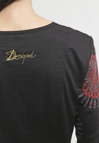 Gros plan d'une chemise noire à manches longues avec un mot brodé en or sur le haut du dos et un motif circulaire rouge sur la manche.