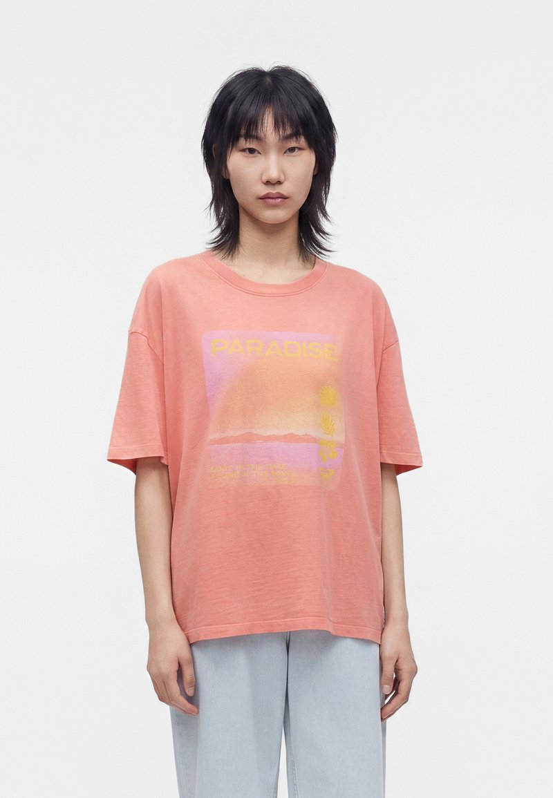 Jeune personne aux cheveux courts et noirs portant un t-shirt corail ample avec le texte "Paradise" et un pantalon bleu clair sur un fond uni.