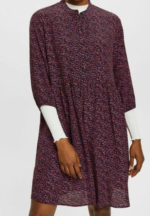 Person trägt ein langärmeliges dunkles Kleid mit kleinem roten, rosa und blauen Blumenmuster über einem weißen, gerippten Langarmshirt mit wellenförmigen Manschetten.