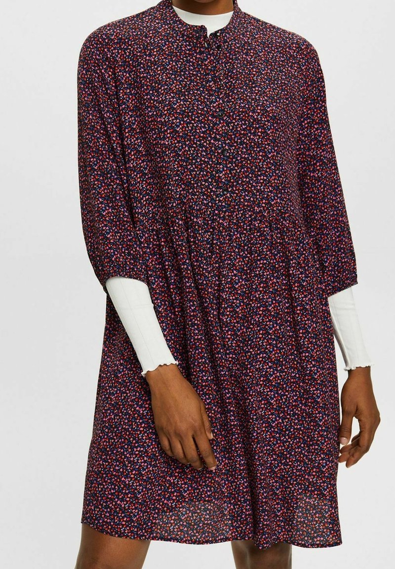 Personne portant une robe longue à manches longues de couleur foncée avec un petit imprimé floral rouge, rose et bleu sur un haut blanc à manches longues en côte avec des poignets festonnés.