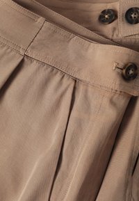 Beige plooibroek met een gladde textuur, voorzien van een brede tailleband, twee donkere knoopsluitingen en gestikte details langs de naden.