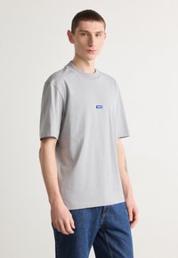Graues Baumwoll-T-Shirt mit kurzen Ärmeln und Rundhalsausschnitt. Mit einem blauen Logo-Aufnäher auf der Brust. Kombiniert mit blauen Jeans.
