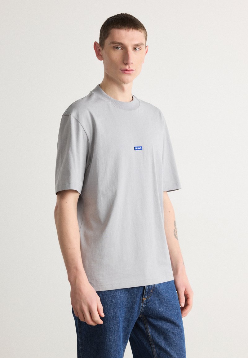 Graues Baumwoll-T-Shirt mit kurzen Ärmeln und Rundhalsausschnitt. Mit einem blauen Logo-Aufnäher auf der Brust. Kombiniert mit blauen Jeans.
