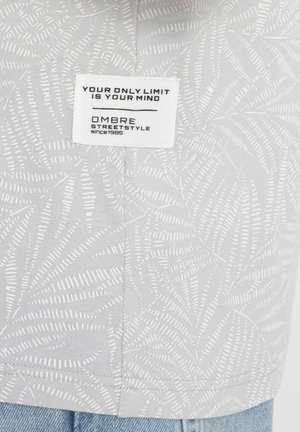 Lichtgrijze stof met witte bladerenpatronen, met een rechthoekig etiket met zwarte tekst die luidt: "JE ENIGE BEPERKING IS JE GEMEENTE OMBARE STREETSTYLE sinds 1985."