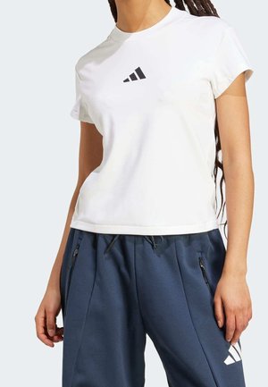 Personne portant un t-shirt blanc Adidas et un pantalon de survêtement Adidas bleu marine, debout avec les mains détendues le long du corps devant un fond uni.