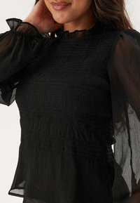 Blusa nera leggera con arricciature testurizzate, maniche a sbuffo trasparenti e dettaglio del colletto arricciato; tessuto morbido con una silhouette fluida.