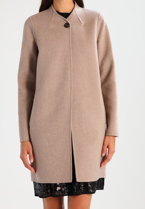Femme portant un long manteau en laine beige avec un bouton noir unique, par-dessus une robe noire à paillettes, debout les mains détendues le long du corps.