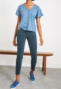 Blå blommig kortärmad blus med V-hals och knapppdetalj, kombinerad med mörkgröna manchesterbyxor och blå sneakers. Naturlig textur.