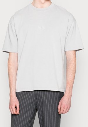 Person trägt ein schlichtes hellgraues T-Shirt mit kurzen Ärmeln und der Aufschrift "YPS" auf der Brust sowie dunkel gestreifte Hose.