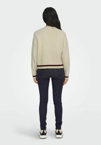 Beżowy sweter dziany z bordowym paskiem u dołu, luźny krój i opadające ramiona, połączony z ciemnymi, obcisłymi dżinsami i czarnymi trampkami.