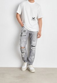 Henkilö, joka käyttää valkoista Calvin Kleinin t-paitaa, harmaita voimakkaasti repaleisia farkkuja ja valkoisia lenkkitossuja seisten tasaista seinää vasten.