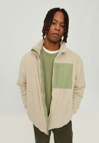 Beige fleece jacket with a green nylon chest pocket, zip closure, and high collar. Layered over a green t-shirt, featuring texture contrast.