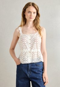 White lace top featuring a buttoned front, ruffled straps, and a tied waist, paired with high-waisted blue denim pants.