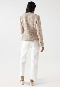 Femme debout de dos, portant une veste en tweed beige, un pantalon blanc taille haute et des sandales blanches à talons blocs sur un fond blanc.