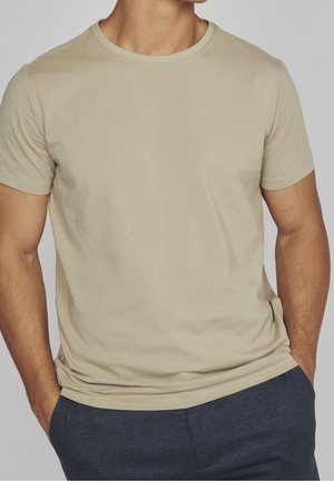 Mężczyzna ubrany w beżowy, krótki rękaw, koszulkę typu crewneck oraz granatowe, w kratę spodnie, ręce w kieszeniach, na jednolitym szarym tle.