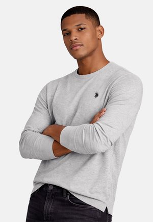 Young man with short hair wearing a light gray long-sleeve shirt and black pants, arms crossed, against a plain white background.