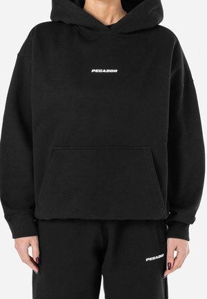 Osoba ubrana w czarną bluzę z kapturem i spodnie z białym logo „PEGADOR” na piersi i udzie, stojąca na jednolitym tle.
