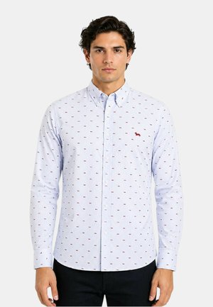 Uomo che indossa una camicia azzurro chiaro con piccolo motivo stampato rosso di cane, in piedi davanti a uno sfondo semplice con le mani lungo i fianchi.