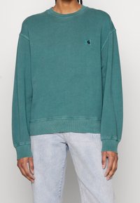 Sudadera de algodón color azul verdoso con cuello redondo, puños y dobladillo acanalados, con un pequeño parche con logo en el pecho izquierdo. Combinada con vaqueros claros.